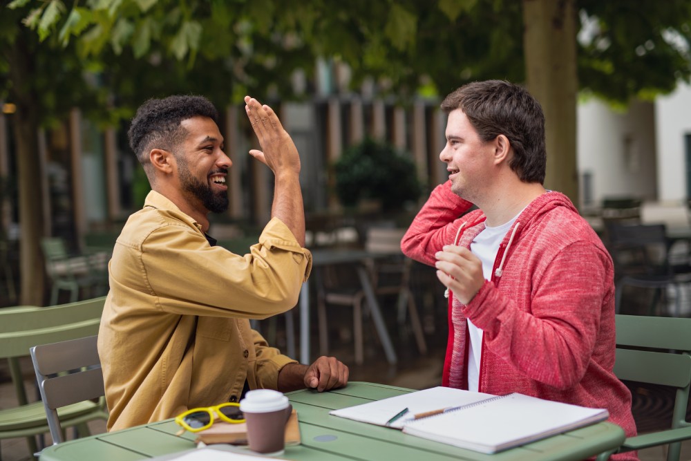 Young man with Down syndrome with mentoring friend sitting outdoors in cafe celebrating success